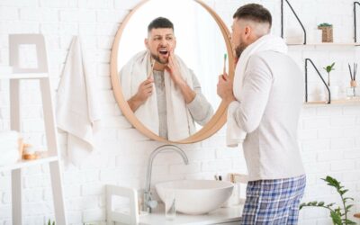 Man holding a toothbrush and looking at his mouth in a bathroom mirror.