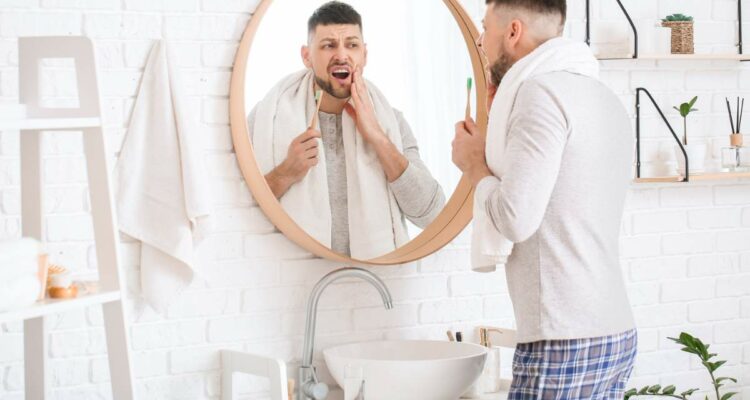 Man holding a toothbrush and looking at his mouth in a bathroom mirror.