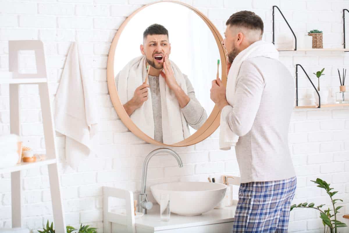 Man holding a toothbrush and looking at his mouth in a bathroom mirror.